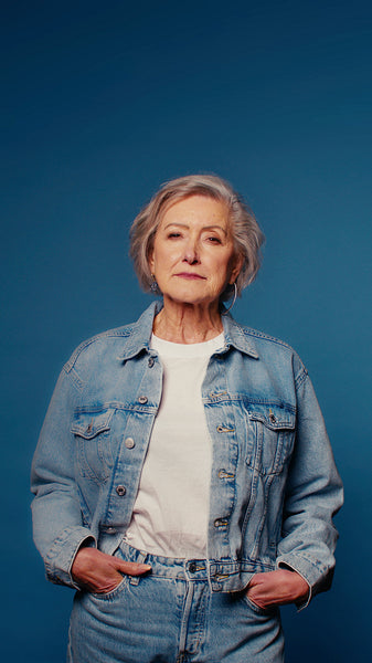 Confident senior woman poses in denim jacket with hands in pockets on a blue background