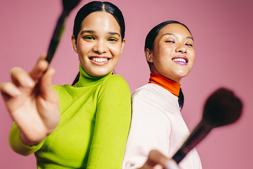 Fun makeup routine, two young women smile at the camera holding their beauty brushes