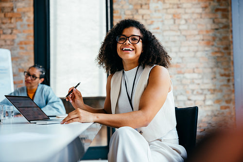 Confident young professional woman smiling in a modern office setting using laptop and pen during a business meeting