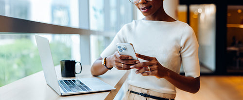 Woman texting on a smartphone while standing near a laptop in an office