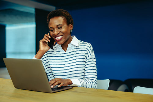 Smiling African businesswoman talking on phone at laptop in office