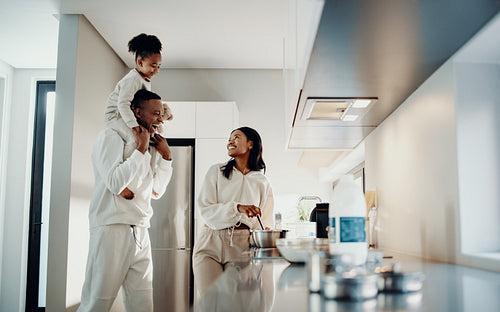 Black family showing parenthood, together in the kitchen - dad carrying daughter, mom cooking