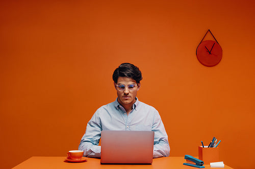 Graphic designer with eyeglasses working on laptop at orange desk