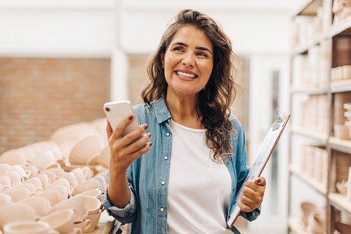 Thoughtful female ceramist holding a smartphone in her shop