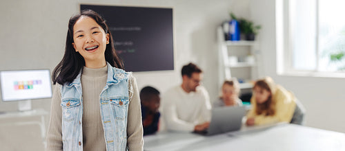 Young Asian student smiling at the camera in a coding class, with her teacher and classmates in the background