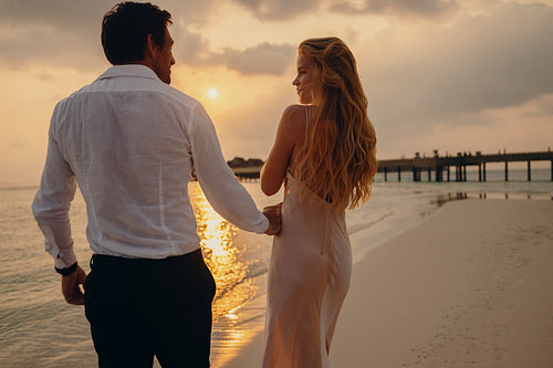 Beautiful blonde woman in gown walking with her partner by the beach at sunset