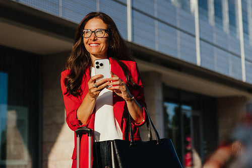 Businesswoman with smartphone outdoors holding luggage and handbag