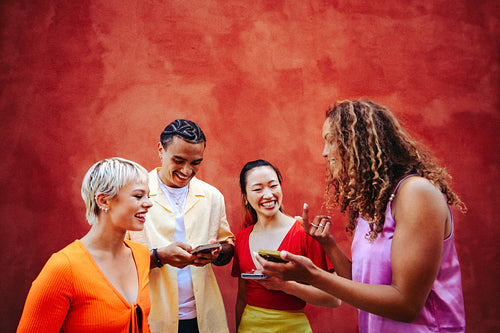 Young adult friends share smartphone screens and laugh together outdoors by a colorful wall