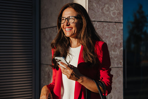 Smiling woman holding phone outdoors wearing a red blazer