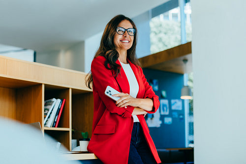 Confident businesswoman in red jacket holding smartphone in bright office environment
