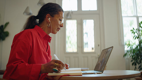 Woman takes a video call on laptop while writing notes in a bright workspace