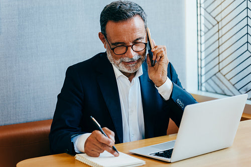 Mature businessman on phone call while noting down details at desk
