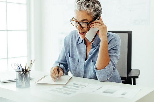 Mature interior designer making notes while speaking to a client on a phone call