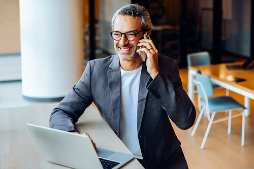 Professional man using a phone while working on a laptop at an office desk