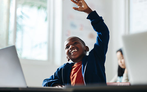 Happy young boy raising his hand to answer a question in a digital literacy classroom