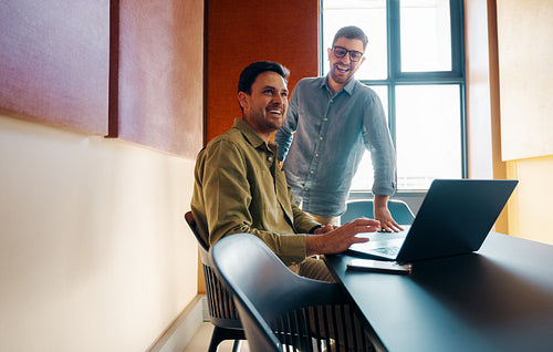 Two mid-adult men laughing and working together at a desk with a laptop