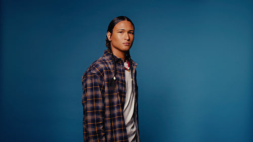 Young man with braids in a plaid shirt, serious gaze, studio portrait on blue