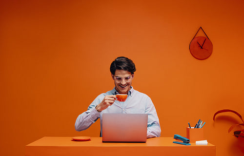 Male entrepreneur drinking coffee while working online in bold orange workspace