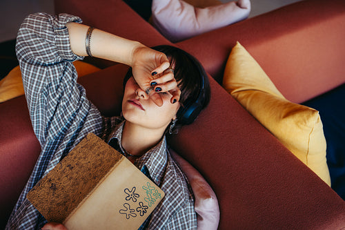 Generation Z individual relaxing with book while listening to music, depicting a peaceful moment of reading and resting on a cozy couch with colorful pillows
