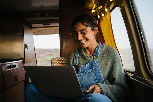 A woman works on a laptop inside a cozy van and smiles at her screen