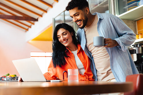Smiling couple enjoying morning coffee while browsing on a laptop in kitchen