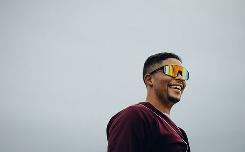 Smiling man wearing sunglasses and baseball attire with a joyful expression on a bright day, exuding confidence and positive energy.
