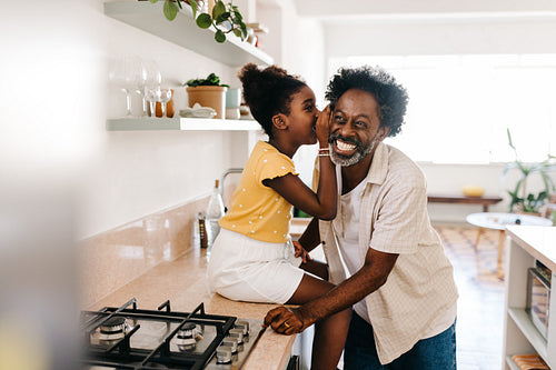 Young girl whispering to her mature dad's ear in the kitchen