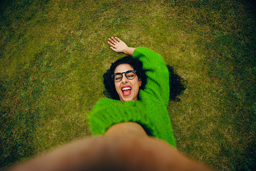 On the grass, the woman is laughing as she takes a selfie outdoors with her smartphone