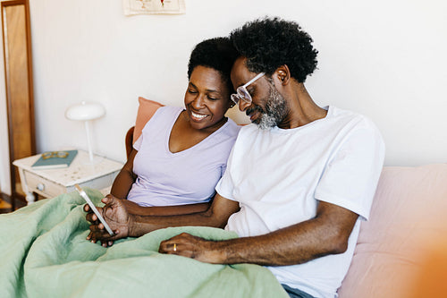 Husband and wife enjoying leisure time together, smiling and browsing social media in a cozy bedroom