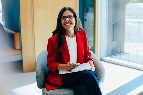 Confident woman wearing red blazer smiling while sitting in a bright office