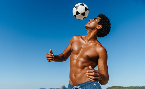 Young man in swim shorts playing soccer on the beach on a sunny summer day