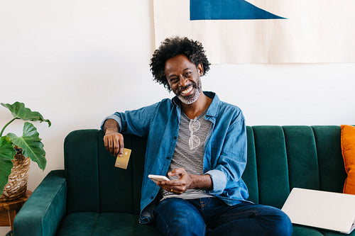 Black man holding a credit card and a phone, using an online shopping app at home