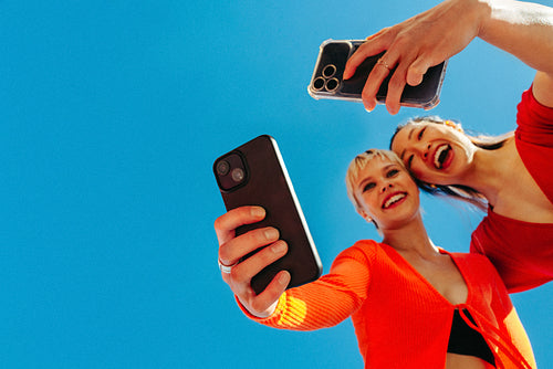 Joyful friends separately taking selfies against a clear blue sky