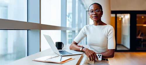 Confident professional woman seated by a window with laptop and notes