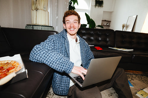 A smiling man uses a laptop in the living room for casual work at home.