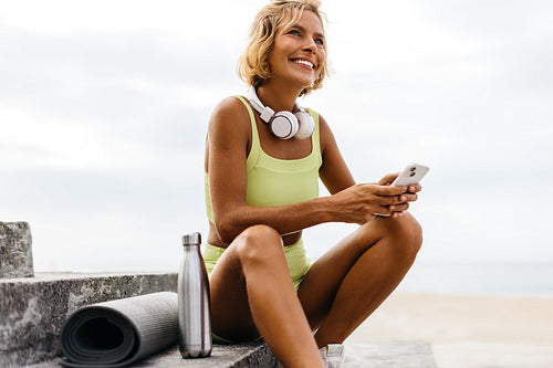 Woman sitting on beachside stairs with her yoga equipment, using her phone to browse a fitness app