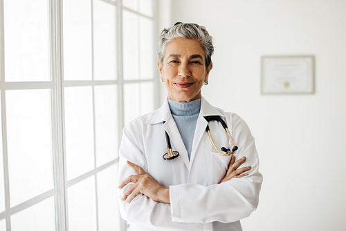Confident in her experience and expertise, mature female doctor stands in her office with her arms crossed