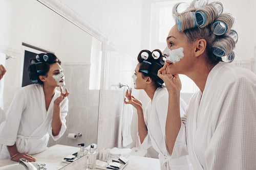 Two women wearing bathrobes applying cream on face standing in front of mirror