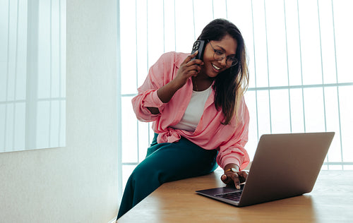 Cheerful businesswoman making a phone call while using a laptop in a modern office