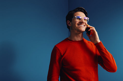 Happy man talking on phone in a colorful colorblocking office setting, wearing orange sweater against blue background