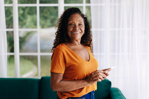 Happy senior woman standing in her living room, holding a smartphone