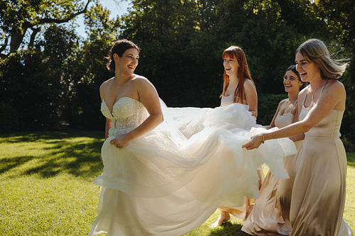 Bride smiling with bridesmaids holding her dress in a sunny garden moment
