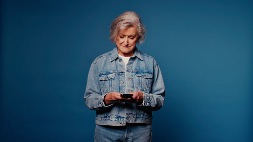 Happy senior woman in denim using smartphone on blue background