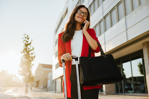 Professional woman with luggage talking on phone outside urban building