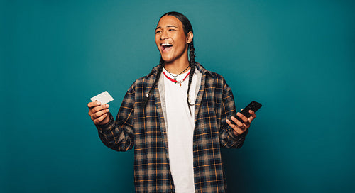 Happy native man smiling with braided hair pays online using credit card on blue studio background