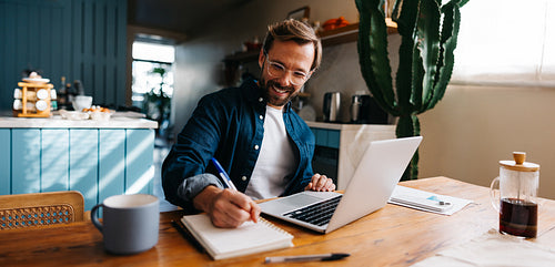Man working at kitchen desk with laptop and notebook