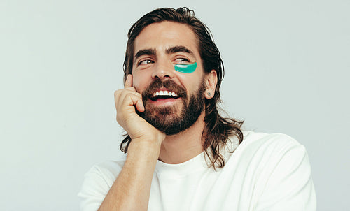 Happy young man caring for his face with an undereye mask in a studio