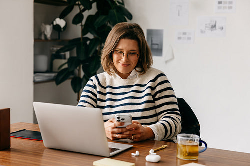 At her desk, the woman balances smartphone tasks with laptop work in a home office
