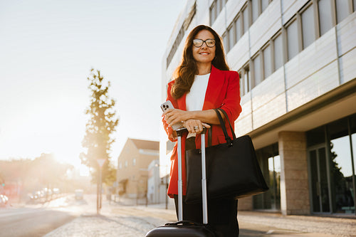 Confident businesswoman outdoors with luggage and mobile phone in urban sunlight