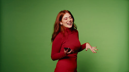Happy redhead woman in red outfit laughs and dances with phone on green screen